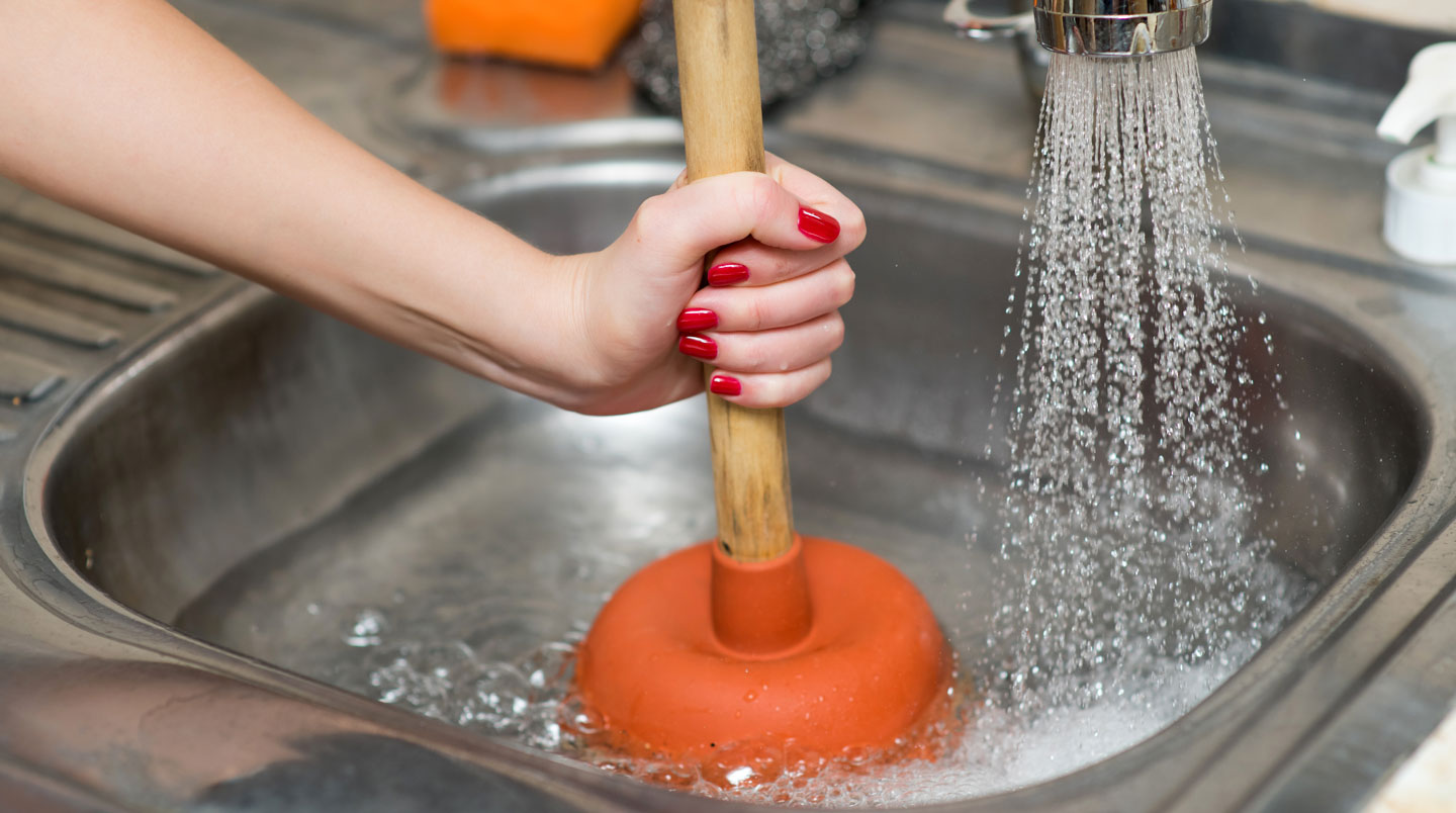 Mains d'un adulte utilisant correctement une ventouse en caoutchouc sur le drain d'un évier de cuisine moderne, avec eau visible recouvrant la coupelle