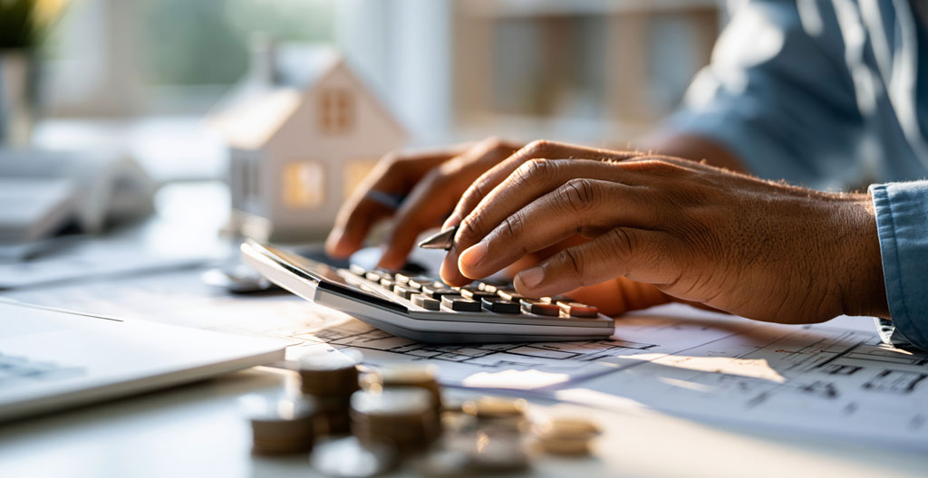Mains d'un particulier consultant plusieurs devis de rénovation et un plan de combles posés sur une table en bois clair avec calculatrice et tablette numérique